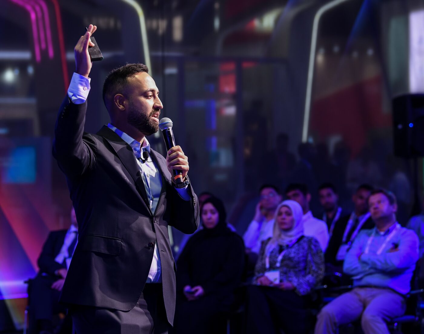Iman Ghanizada delivering a keynote, microphone in hand, on a darkened stage.